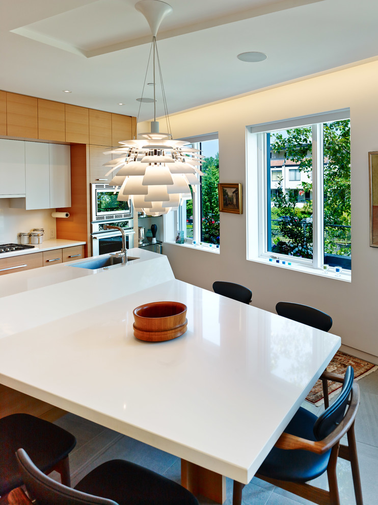 Стильный дизайн: прямая кухня в стиле модернизм с обеденным столом, врезной мойкой, плоскими фасадами, светлыми деревянными фасадами, мраморной столешницей, белым фартуком и техникой из нержавеющей стали - последний тренд