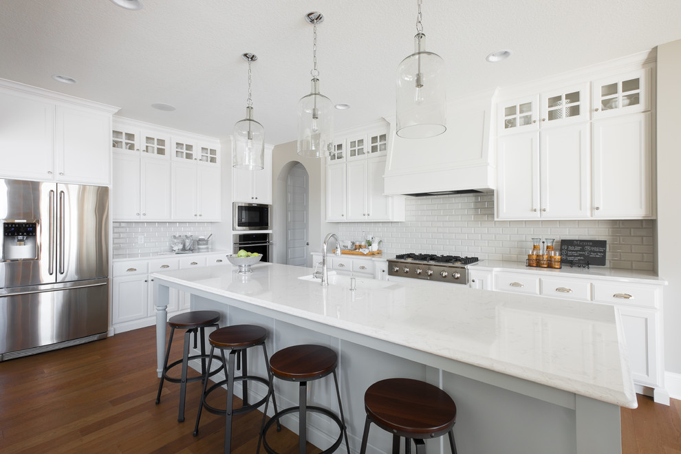 Elegant l-shaped medium tone wood floor kitchen photo in Minneapolis with shaker cabinets, white cabinets, white backsplash, subway tile backsplash, stainless steel appliances, an island, a farmhouse sink and quartz countertops