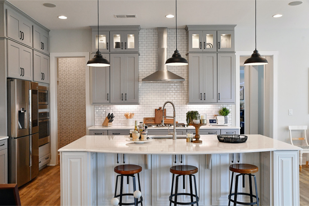 Example of a transitional l-shaped medium tone wood floor and brown floor kitchen design in Charlotte with an undermount sink, shaker cabinets, gray cabinets, white backsplash, subway tile backsplash, stainless steel appliances, an island and white countertops