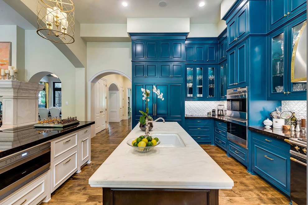 Große Moderne Wohnküche in L-Form mit blauen Schränken, Küchenrückwand in Weiß, braunem Holzboden, Landhausspüle, Schrankfronten mit vertiefter Füllung, Küchengeräten aus Edelstahl, zwei Kücheninseln, braunem Boden und schwarzer Arbeitsplatte in Dallas