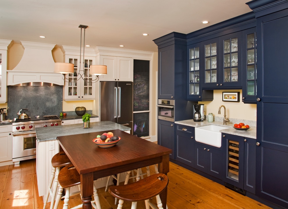 На фото: п-образная кухня в стиле кантри с обеденным столом, с полувстраиваемой мойкой (с передним бортиком), стеклянными фасадами, синими фасадами, серым фартуком и техникой из нержавеющей стали с