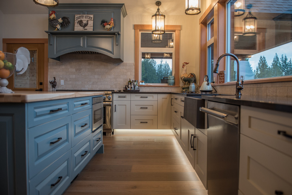 Example of a large farmhouse u-shaped light wood floor and brown floor kitchen design in Other with a farmhouse sink, recessed-panel cabinets, distressed cabinets, quartzite countertops, beige backsplash, limestone backsplash, stainless steel appliances, an island and black countertops