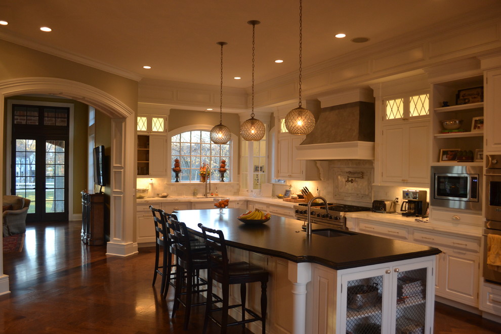 Klassisk inredning av ett stort kök, med en undermonterad diskho, luckor med profilerade fronter, vita skåp, grått stänkskydd, stänkskydd i stenkakel, rostfria vitvaror, mellanmörkt trägolv och en köksö