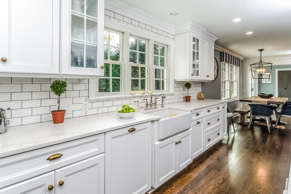Example of a large classic l-shaped dark wood floor and brown floor eat-in kitchen design in New York with a farmhouse sink, recessed-panel cabinets, white cabinets, marble countertops, white backsplash, subway tile backsplash, stainless steel appliances and an island