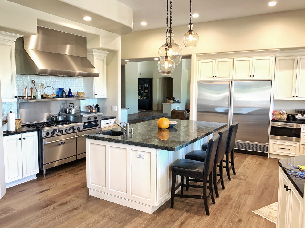 Eat-in kitchen - huge transitional u-shaped light wood floor and beige floor eat-in kitchen idea in Phoenix with a farmhouse sink, flat-panel cabinets, white cabinets, quartz countertops, white backsplash, glass tile backsplash, stainless steel appliances, an island and black countertops