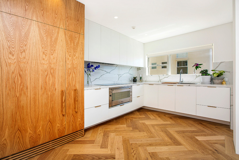 Modern inredning av ett kök, med en nedsänkt diskho, släta luckor, vita skåp, vitt stänkskydd, rostfria vitvaror och ljust trägolv