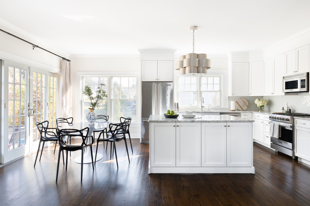 Photo of a classic l-shaped kitchen/diner in New York with shaker cabinets, white cabinets, grey splashback, stainless steel appliances, dark hardwood flooring, an island, brown floors and grey worktops.