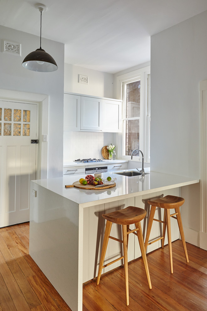 This is an example of a medium sized classic u-shaped kitchen/diner in Sydney with a built-in sink, shaker cabinets, white cabinets, engineered stone countertops, white splashback, ceramic splashback, white appliances, light hardwood flooring and an island.