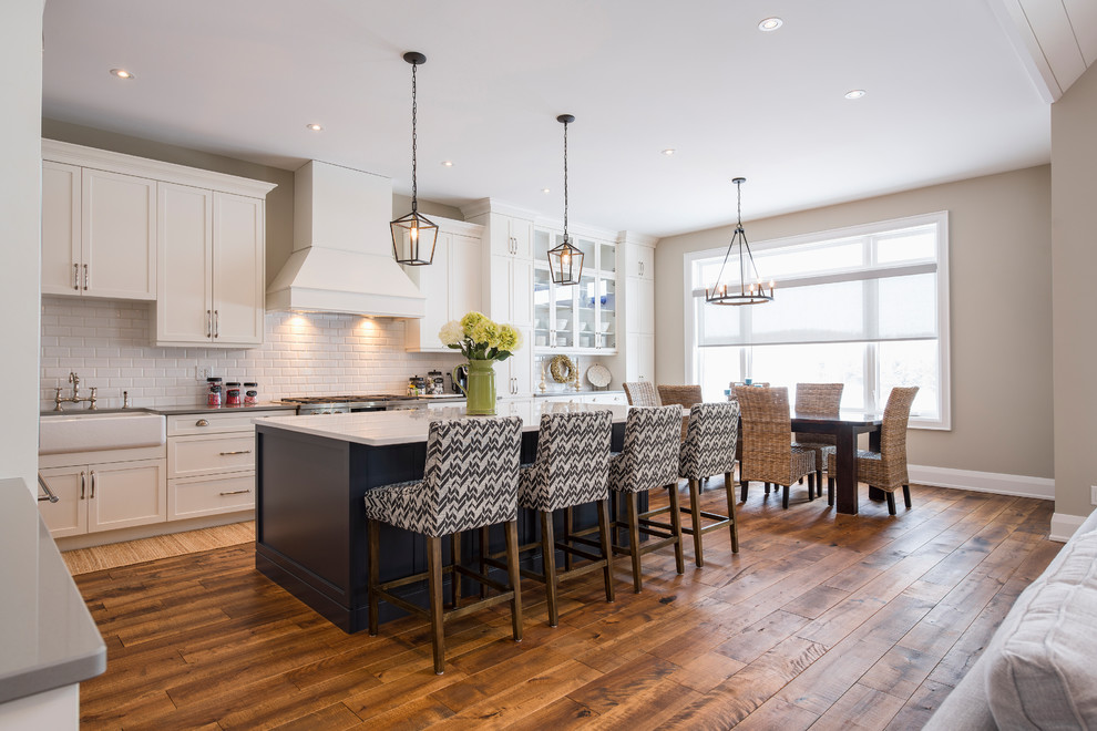 Example of a large transitional l-shaped medium tone wood floor and brown floor eat-in kitchen design in Toronto with a farmhouse sink, shaker cabinets, white cabinets, white backsplash, porcelain backsplash, stainless steel appliances and an island