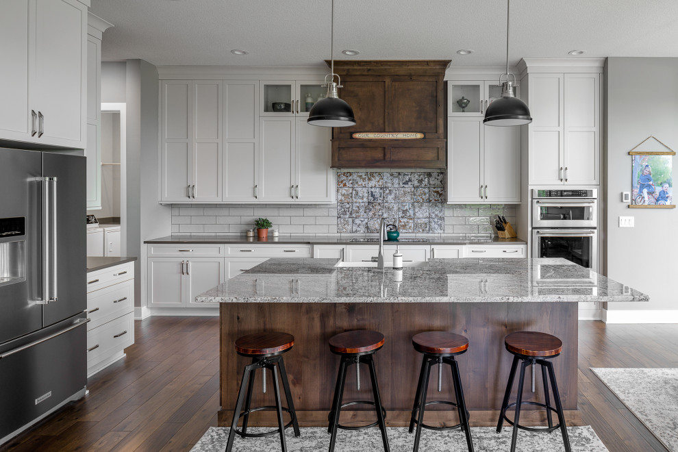 Country l-shaped dark wood floor and brown floor kitchen photo in Other with a farmhouse sink, shaker cabinets, white cabinets, white backsplash, stainless steel appliances, an island and gray countertops
