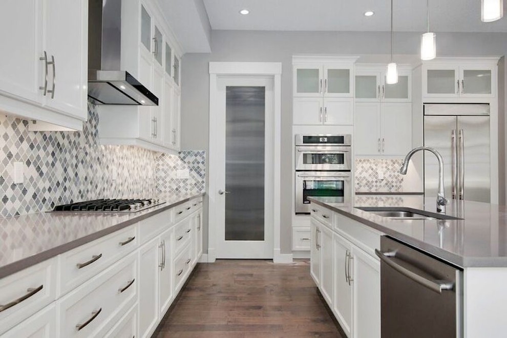Large l-shaped kitchen/diner in Calgary with a double-bowl sink, shaker cabinets, white cabinets, composite countertops, grey splashback, mosaic tiled splashback, stainless steel appliances, medium hardwood flooring and an island.