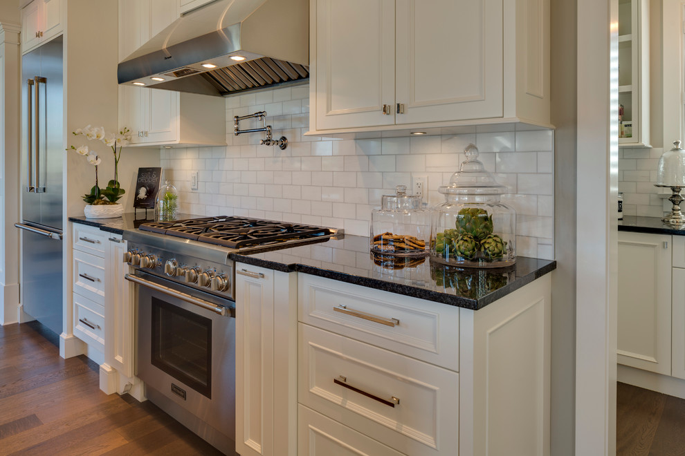 Klassisk inredning av ett mellanstort kök, med luckor med infälld panel, vita skåp, granitbänkskiva, stänkskydd i tunnelbanekakel, rostfria vitvaror, mellanmörkt trägolv och en köksö