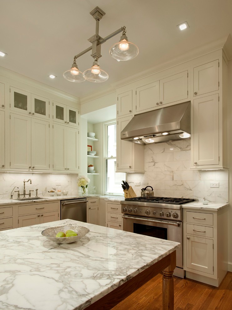 Elegant medium tone wood floor kitchen photo in New York with an undermount sink, recessed-panel cabinets, white cabinets, marble countertops, stainless steel appliances, white backsplash and an island