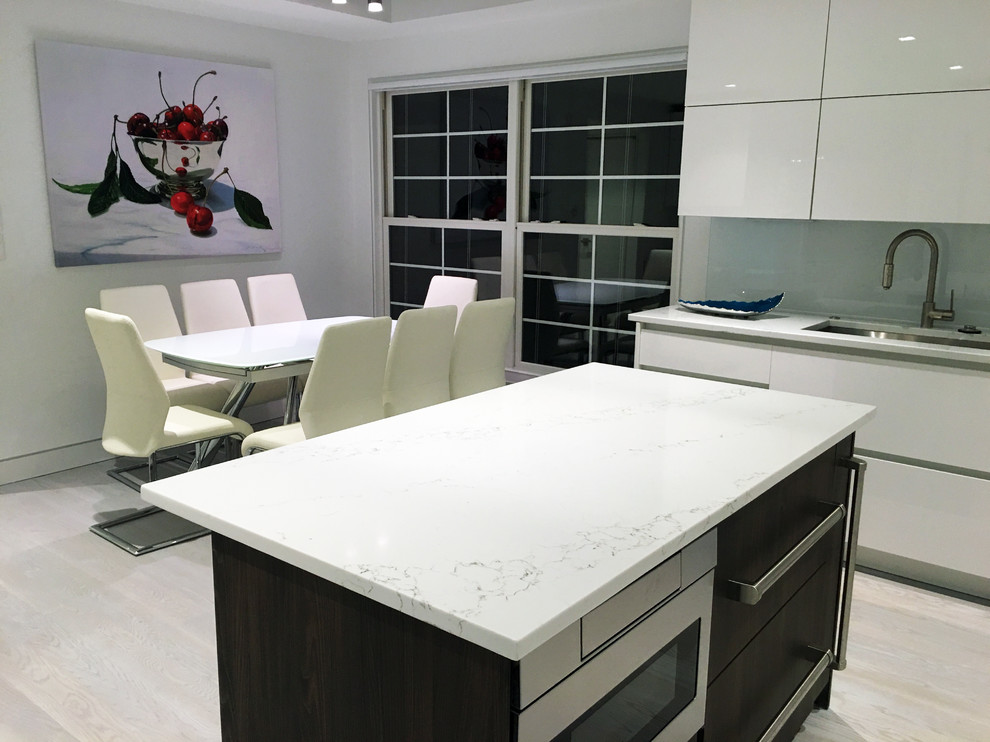 This is an example of a large modern u-shaped open plan kitchen in New York with a built-in sink, flat-panel cabinets, white cabinets, marble worktops, white splashback, glass sheet splashback, black appliances, light hardwood flooring and an island.