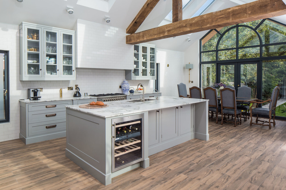 Foto de cocina comedor lineal clásica renovada grande con encimera de mármol, salpicadero blanco, electrodomésticos de acero inoxidable, una isla, puertas de armario grises, salpicadero de azulejos tipo metro, fregadero de doble seno, armarios estilo shaker y suelo de madera oscura