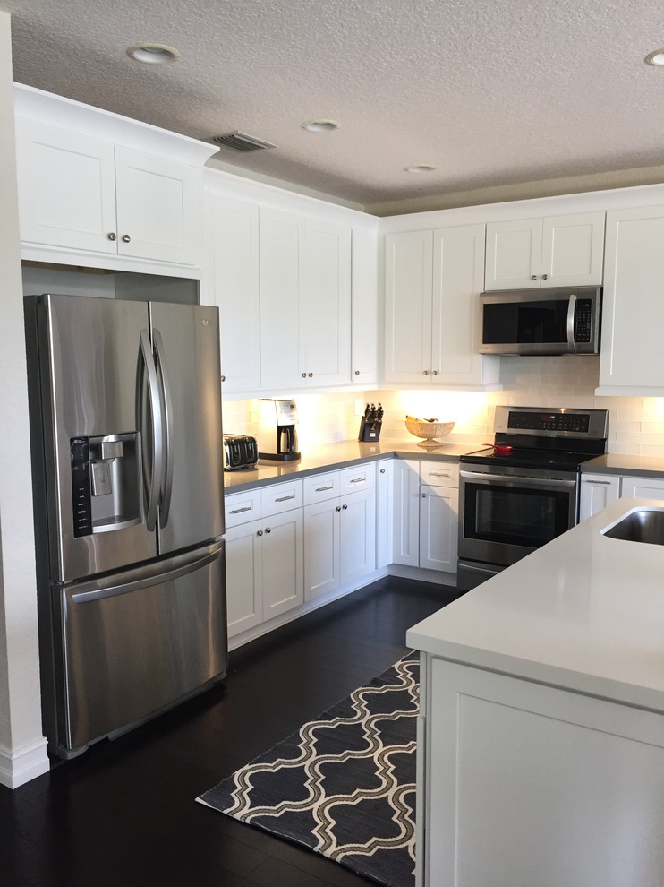 Photo of a medium sized contemporary l-shaped kitchen/diner in Miami with a single-bowl sink, shaker cabinets, white cabinets, engineered stone countertops, white splashback, glass tiled splashback, stainless steel appliances, dark hardwood flooring and an island.