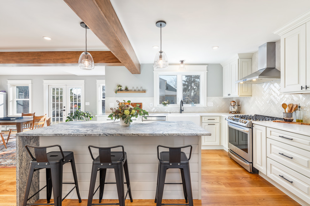 Photo of a medium sized classic l-shaped kitchen/diner in Boston with a belfast sink, raised-panel cabinets, white cabinets, engineered stone countertops, white splashback, metro tiled splashback, stainless steel appliances, light hardwood flooring, an island, beige floors and white worktops.