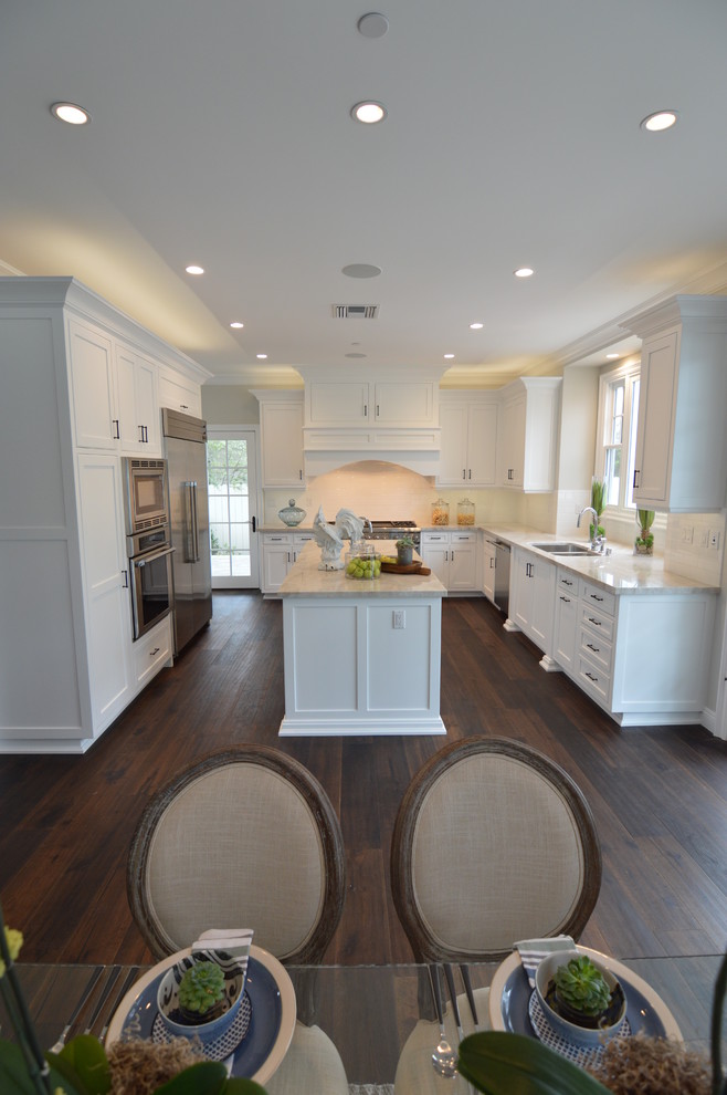 Bild på ett stort funkis kök, med en dubbel diskho, luckor med infälld panel, vita skåp, marmorbänkskiva, vitt stänkskydd, rostfria vitvaror, mellanmörkt trägolv, en köksö, brunt golv och stänkskydd i mosaik