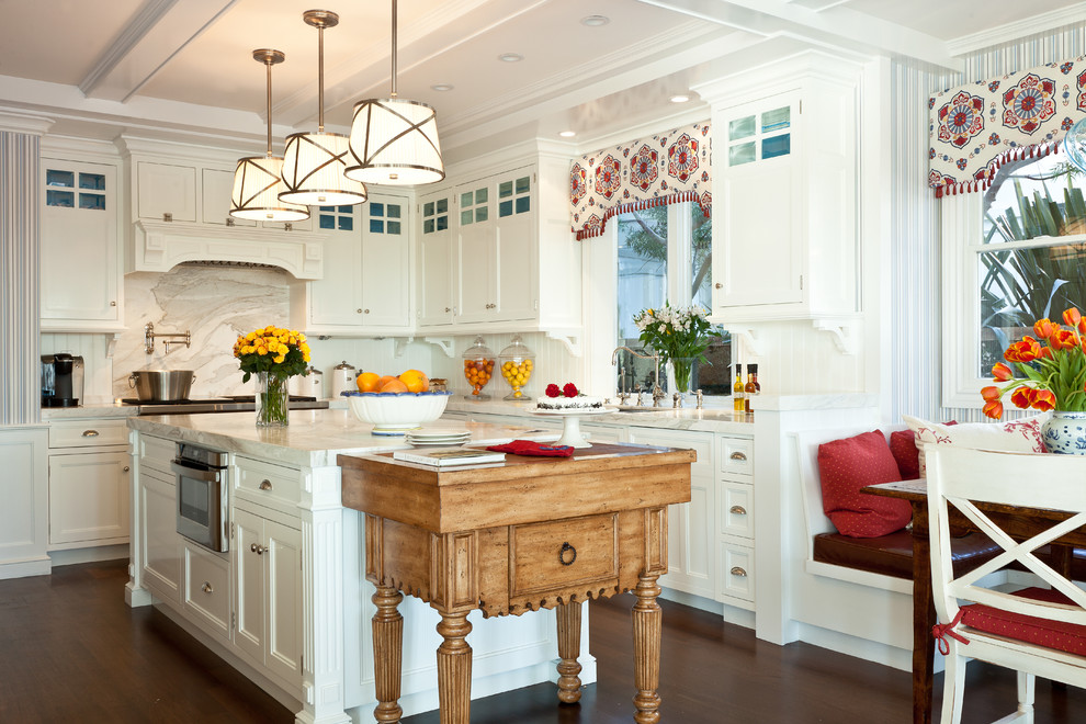 Inredning av ett klassiskt mellanstort vit vitt kök, med en undermonterad diskho, luckor med infälld panel, vita skåp, flerfärgad stänkskydd, rostfria vitvaror, mellanmörkt trägolv, flera köksöar, marmorbänkskiva, stänkskydd i trä och brunt golv