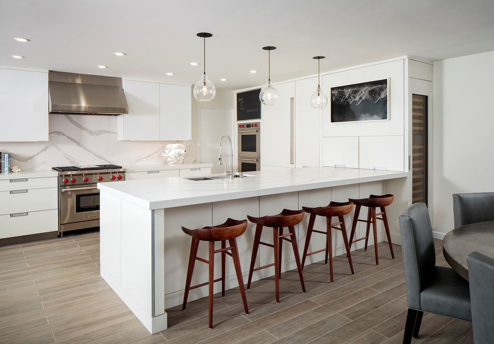 Contemporary u-shaped kitchen in Orange County with a submerged sink, flat-panel cabinets, white cabinets, white splashback, stainless steel appliances, a breakfast bar, brown floors and white worktops.