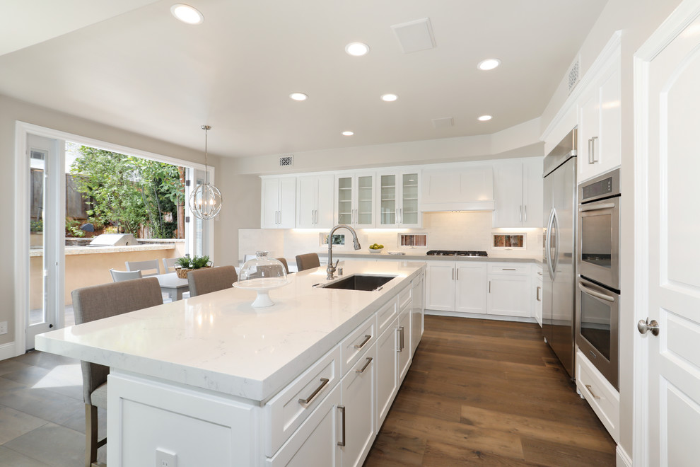 Photo of a medium sized classic grey and white l-shaped kitchen/diner in Orange County with a submerged sink, shaker cabinets, white cabinets, quartz worktops, white splashback, mosaic tiled splashback, stainless steel appliances, an island, brown floors, medium hardwood flooring and white worktops.