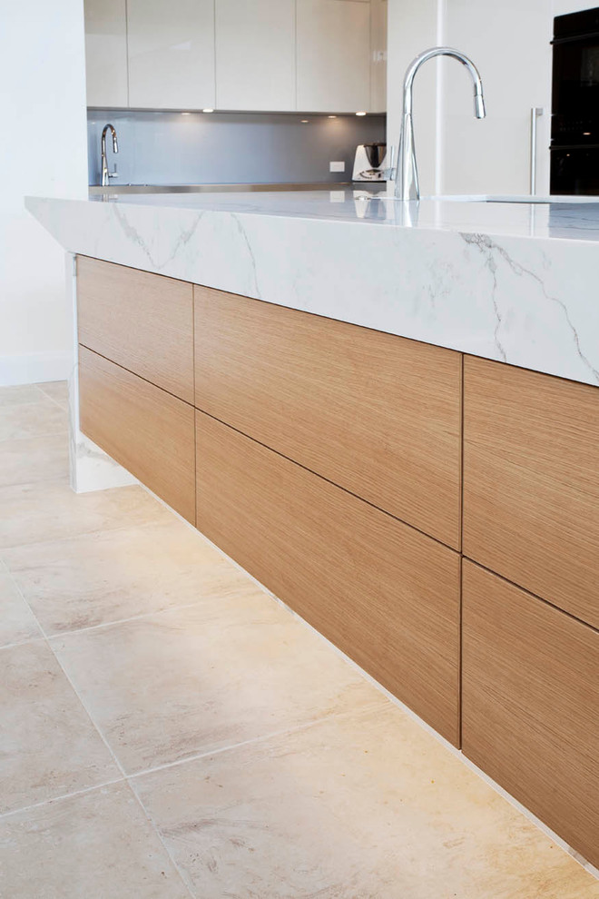 Inspiration for a large modern l-shaped open plan kitchen in Sydney with a submerged sink, flat-panel cabinets, white cabinets, engineered stone countertops, grey splashback, black appliances, limestone flooring, an island and glass sheet splashback.