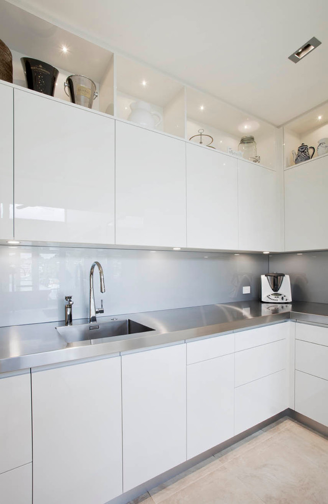 Foto på ett stort funkis kök, med en undermonterad diskho, släta luckor, vita skåp, bänkskiva i kvarts, grått stänkskydd, spegel som stänkskydd, svarta vitvaror, kalkstensgolv och en köksö