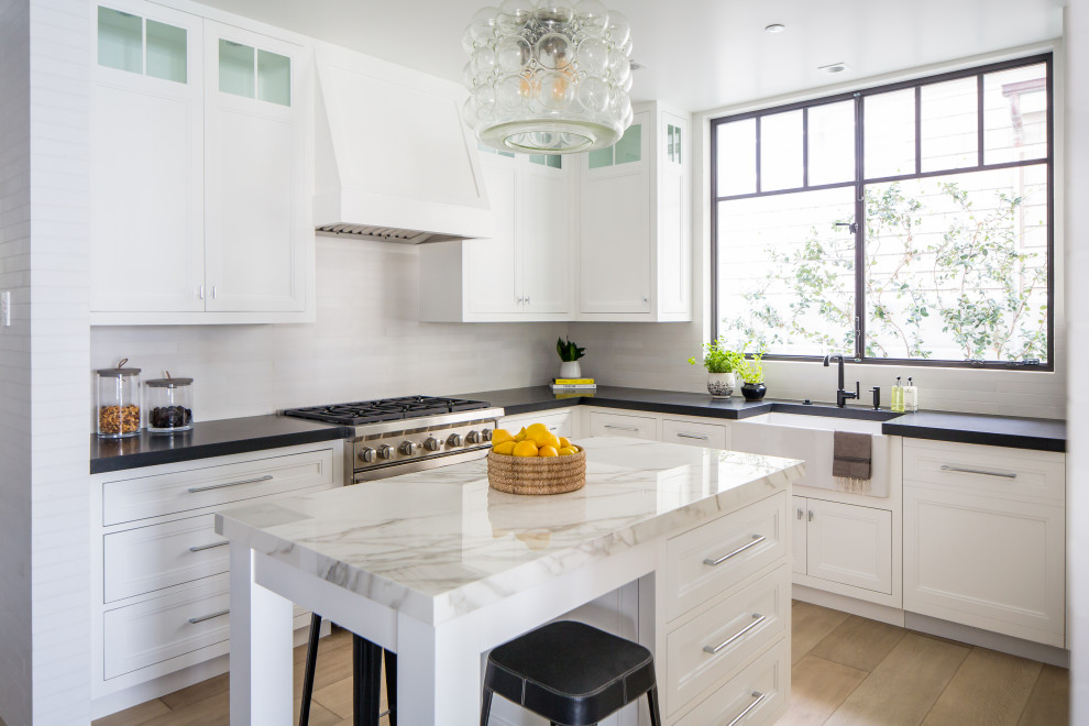 Inspiration for a mid-sized coastal l-shaped light wood floor and beige floor kitchen remodel in Orange County with a farmhouse sink, white cabinets, stainless steel appliances, an island, shaker cabinets, white backsplash and black countertops