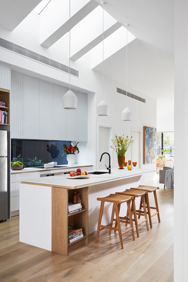 Идея дизайна: кухня-гостиная в современном стиле с одинарной мойкой, белыми фасадами, синим фартуком, фартуком из стекла, техникой из нержавеющей стали, светлым паркетным полом, островом, бежевым полом и белой столешницей