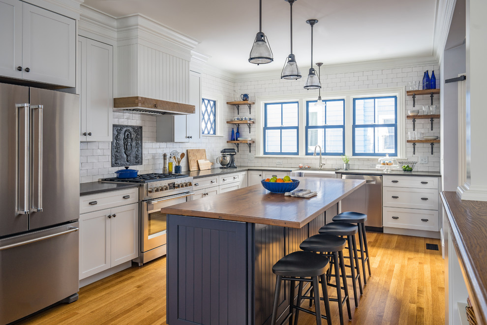 Photo of a farmhouse l-shaped kitchen/diner in Boston with a belfast sink, shaker cabinets, white cabinets, wood worktops, white splashback, metro tiled splashback, stainless steel appliances, medium hardwood flooring, an island and brown floors.