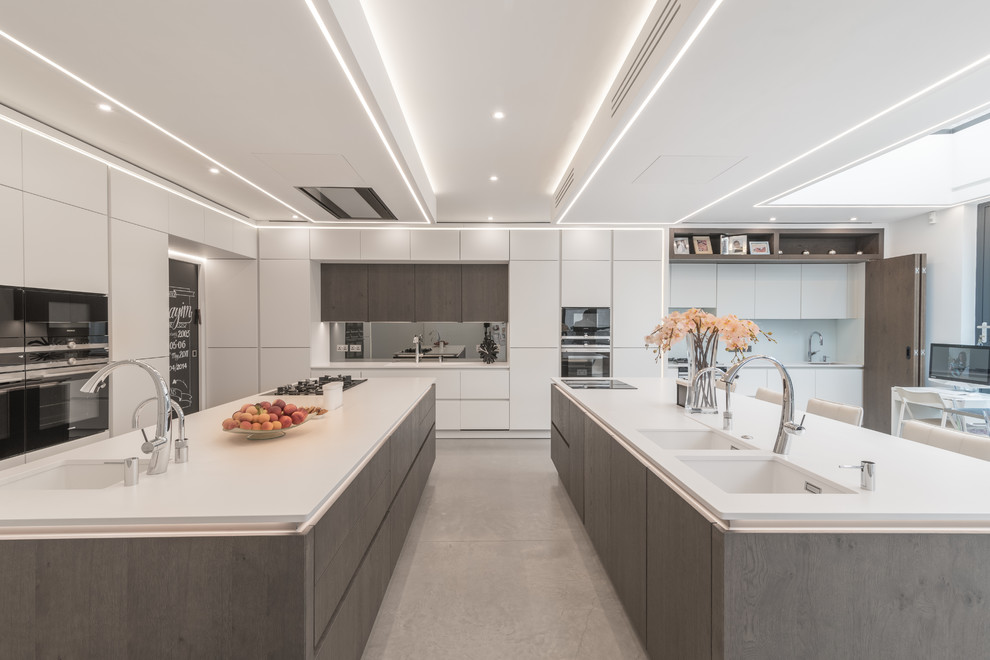 This is an example of a contemporary l-shaped kitchen/diner in London with a submerged sink, flat-panel cabinets, white cabinets, metallic splashback, mirror splashback, black appliances, multiple islands, grey floors and white worktops.