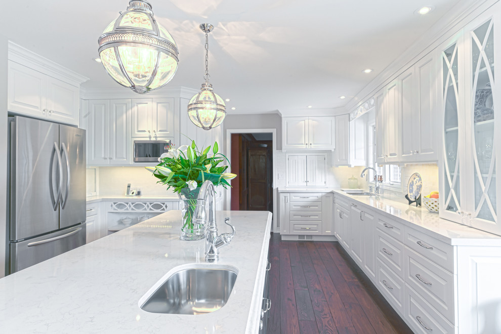 Inredning av ett modernt stort kök, med en köksö, luckor med profilerade fronter, vita skåp, granitbänkskiva, vitt stänkskydd, stänkskydd i keramik, rostfria vitvaror, en nedsänkt diskho och mellanmörkt trägolv