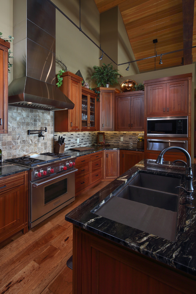 Foto på ett stort amerikanskt kök, med en undermonterad diskho, luckor med infälld panel, skåp i mellenmörkt trä, granitbänkskiva, brunt stänkskydd, stänkskydd i keramik, rostfria vitvaror, ljust trägolv, flera köksöar och brunt golv