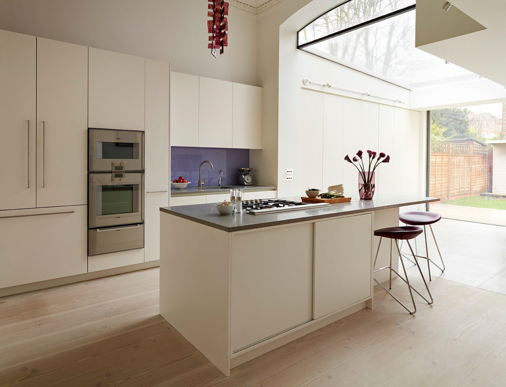 Design ideas for a large contemporary single-wall open plan kitchen in London with flat-panel cabinets, white cabinets, quartz worktops, glass tiled splashback, stainless steel appliances, light hardwood flooring, an island, beige floors and brown worktops.