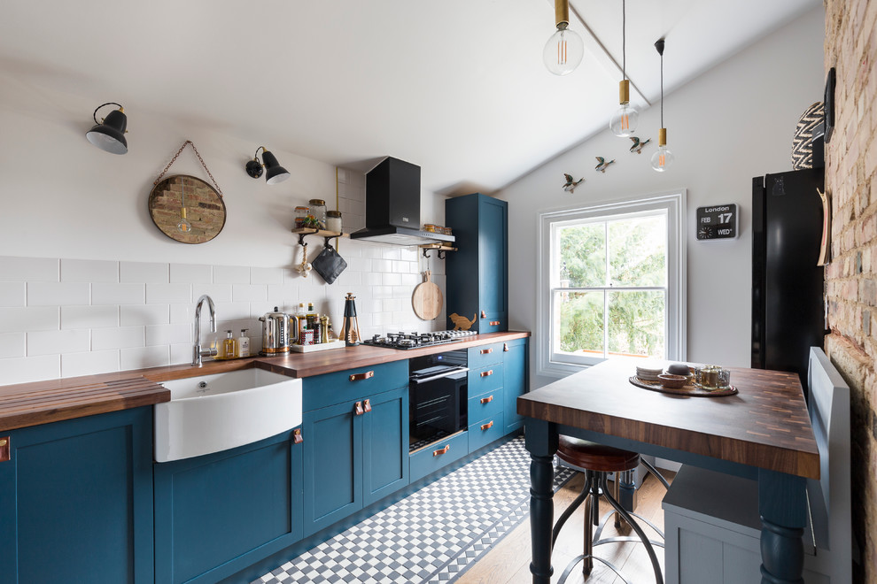Kitchen - contemporary galley kitchen idea in London with a farmhouse sink, shaker cabinets, blue cabinets, wood countertops, white backsplash, subway tile backsplash, black appliances and an island
