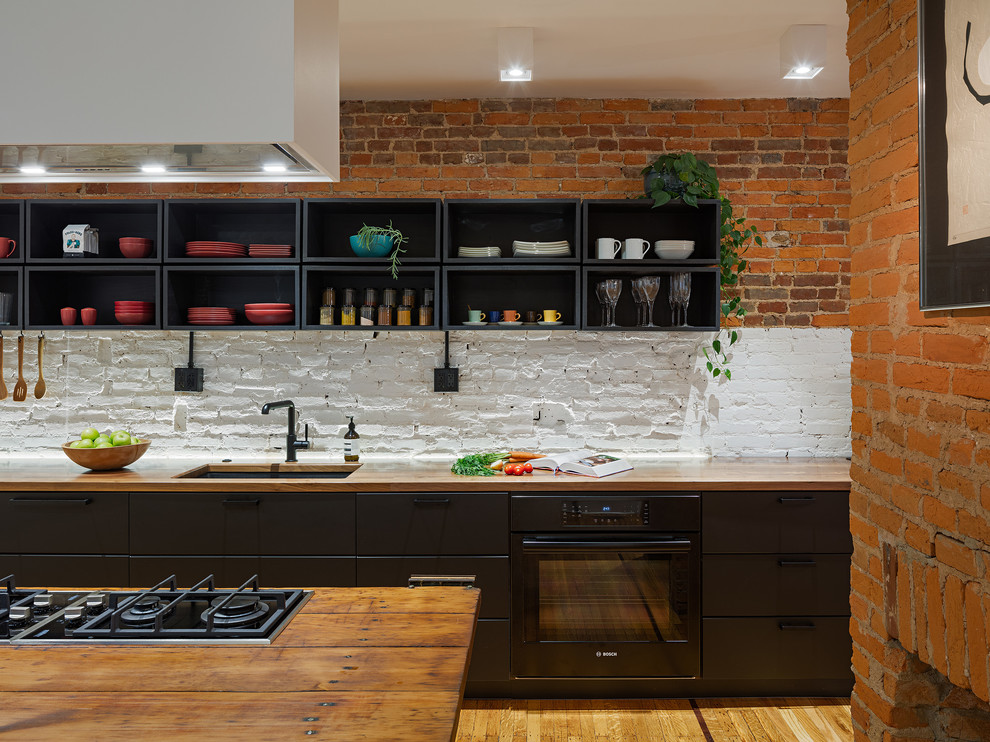 Inspiration for a mid-sized scandinavian medium tone wood floor and brown floor eat-in kitchen remodel in Philadelphia with an undermount sink, flat-panel cabinets, black cabinets, wood countertops, white backsplash, brick backsplash, black appliances, an island and brown countertops