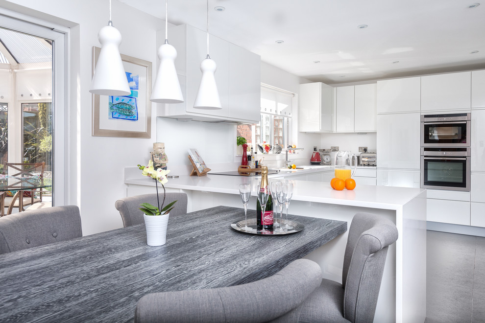 Moderne Wohnküche in U-Form mit flächenbündigen Schrankfronten, Küchengeräten aus Edelstahl, weißen Schränken, Glasrückwand, Küchenrückwand in Weiß, Einbauwaschbecken und Quarzit-Arbeitsplatte in Devon