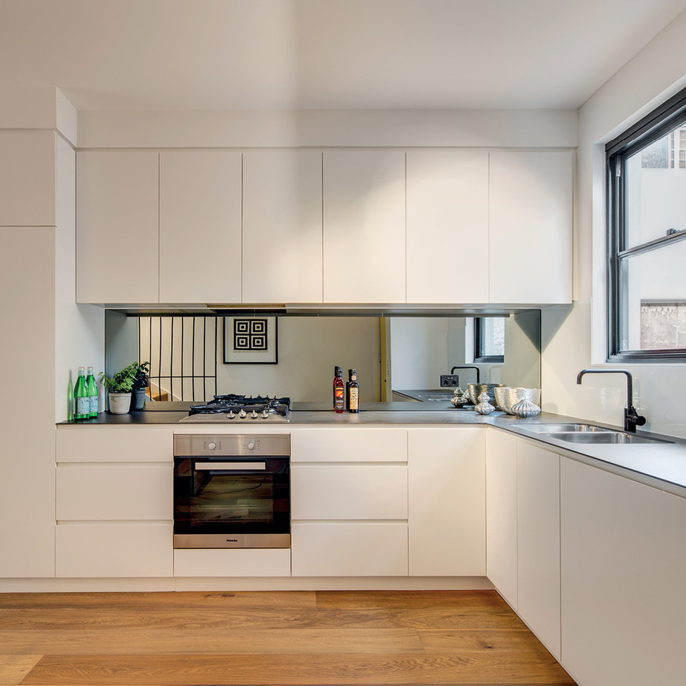 Источник вдохновения для домашнего уюта: угловая кухня в современном стиле с белыми фасадами, фартуком цвета металлик, зеркальным фартуком и светлым паркетным полом без острова