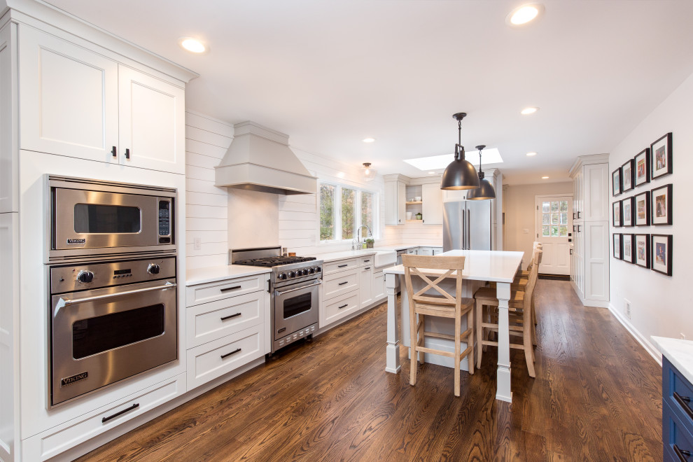 Inredning av ett lantligt stort vit vitt kök, med en rustik diskho, skåp i shakerstil, grå skåp, bänkskiva i kvarts, vitt stänkskydd, stänkskydd i trä, rostfria vitvaror, mellanmörkt trägolv, en köksö och brunt golv