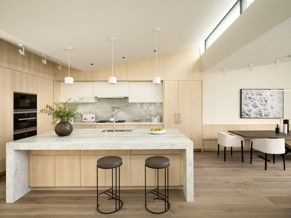 Bild på ett funkis vit vitt l-kök, med en undermonterad diskho, släta luckor, skåp i ljust trä, grått stänkskydd, stänkskydd i sten, rostfria vitvaror, mellanmörkt trägolv, en köksö och brunt golv