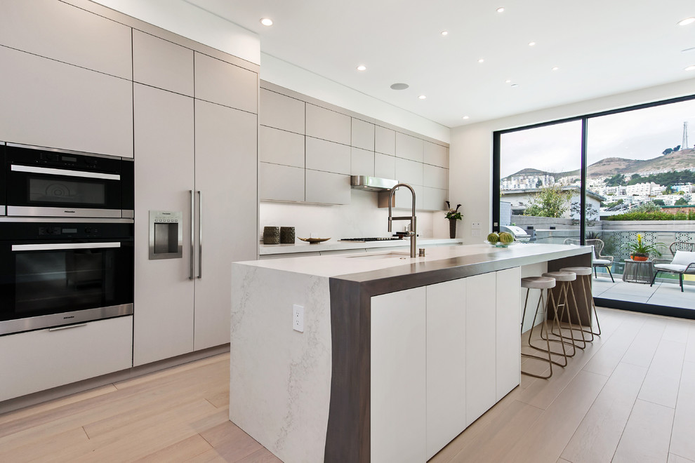Photo of a contemporary galley kitchen in San Francisco with a submerged sink, flat-panel cabinets, beige cabinets, white splashback, integrated appliances, light hardwood flooring, an island and beige floors.
