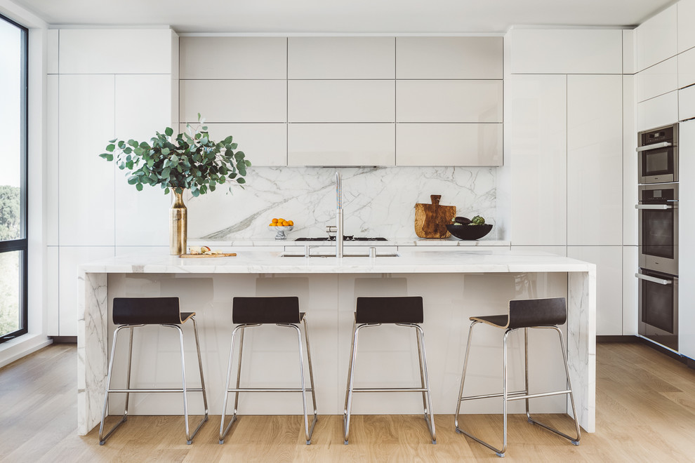 This is an example of a contemporary kitchen in San Francisco with a submerged sink, flat-panel cabinets, white cabinets, white splashback, stone slab splashback, stainless steel appliances, light hardwood flooring and an island.