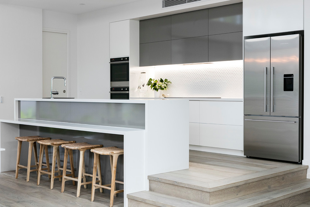 This is an example of a beach style galley kitchen in Sunshine Coast with flat-panel cabinets, white cabinets, white splashback, stainless steel appliances, light hardwood flooring, a breakfast bar, beige floors and white worktops.