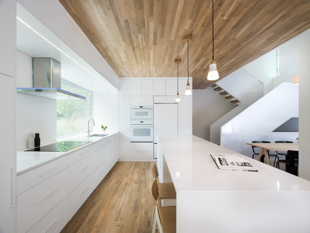 This is an example of a medium sized modern l-shaped kitchen/diner in Other with a built-in sink, flat-panel cabinets, white cabinets, white splashback, white appliances, light hardwood flooring, an island and brown floors.
