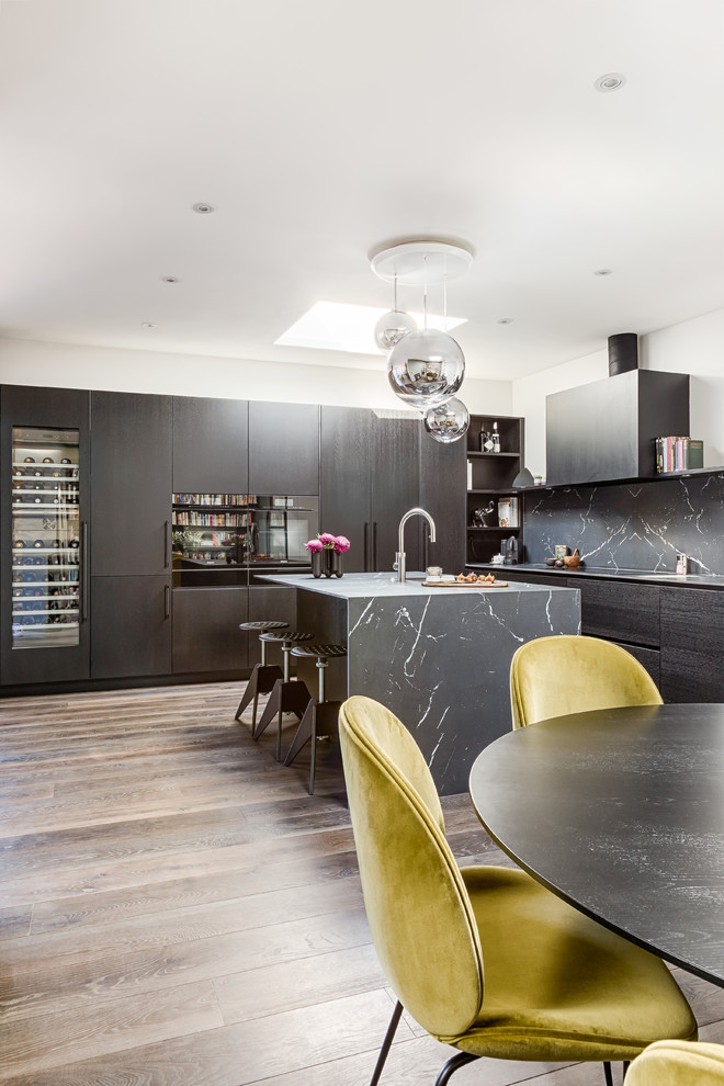This is an example of a medium sized industrial l-shaped open plan kitchen in London with an integrated sink, flat-panel cabinets, black cabinets, composite countertops, black splashback, stone slab splashback, black appliances, medium hardwood flooring, an island, beige floors and black worktops.