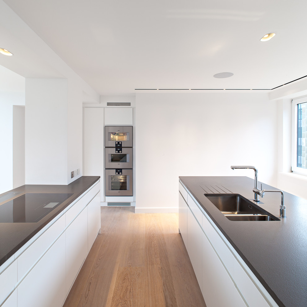 Cette image montre une cuisine design avec un évier 2 bacs, un placard à porte plane, des portes de placard blanches, un électroménager en acier inoxydable, parquet clair et 2 îlots.