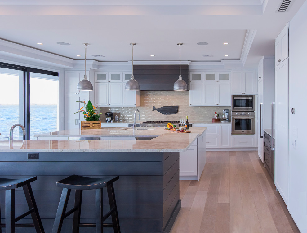 Photo of a nautical l-shaped kitchen in New York with a submerged sink, shaker cabinets, white cabinets, beige splashback, stainless steel appliances, light hardwood flooring, multiple islands, beige floors and beige worktops.