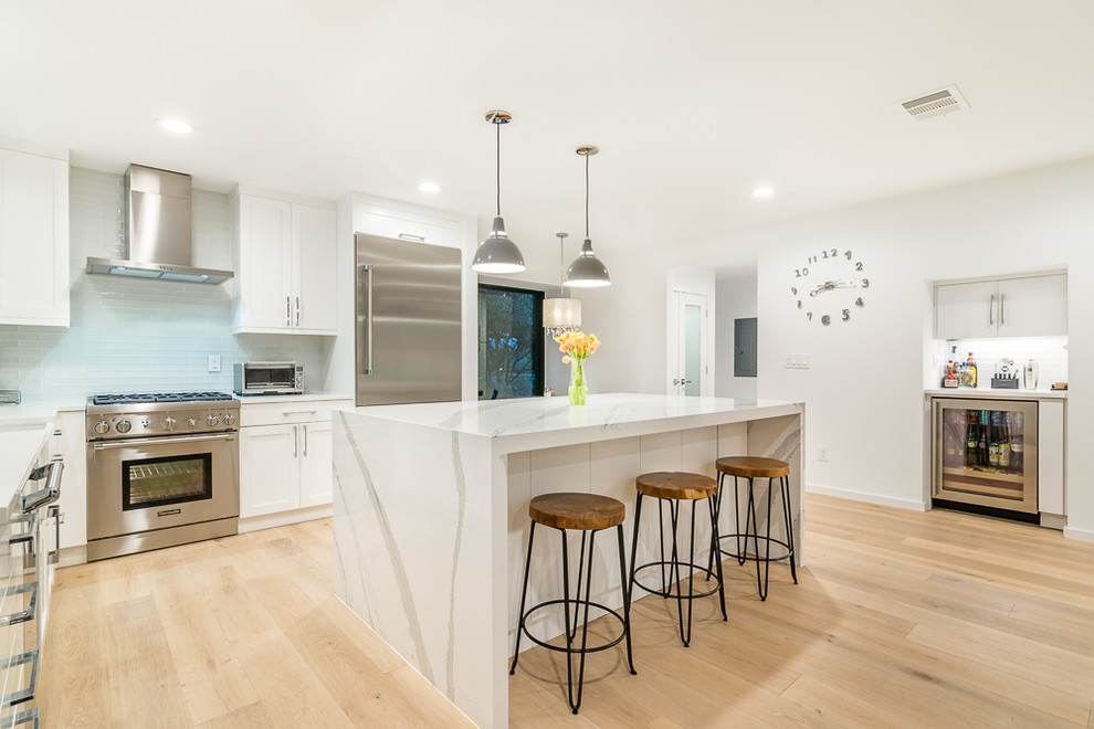 Example of a mid-sized minimalist l-shaped light wood floor open concept kitchen design in Austin with a farmhouse sink, shaker cabinets, white cabinets, white backsplash, subway tile backsplash, stainless steel appliances and an island