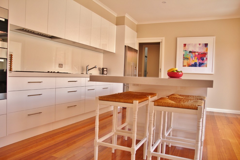 Zweizeilige, Mittelgroße Moderne Wohnküche mit Doppelwaschbecken, flächenbündigen Schrankfronten, weißen Schränken, Quarzwerkstein-Arbeitsplatte, Küchenrückwand in Weiß, Glasrückwand, Küchengeräten aus Edelstahl, braunem Holzboden und Kücheninsel in Melbourne