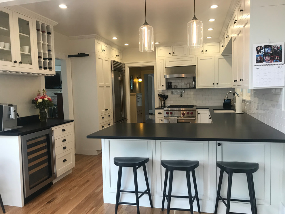 Example of a mid-sized classic u-shaped medium tone wood floor enclosed kitchen design in Seattle with an undermount sink, shaker cabinets, white cabinets, granite countertops, white backsplash, ceramic backsplash, stainless steel appliances, a peninsula and black countertops